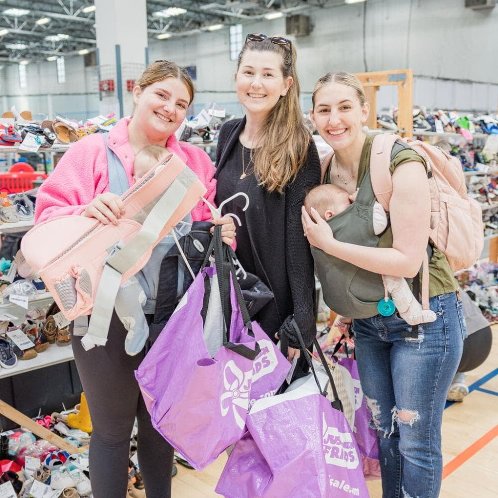 Three women, one with a baby in an infant carrier on her chest, smile while holding purple shopping bags, kid clothing, and a pink toddler hip carrier.