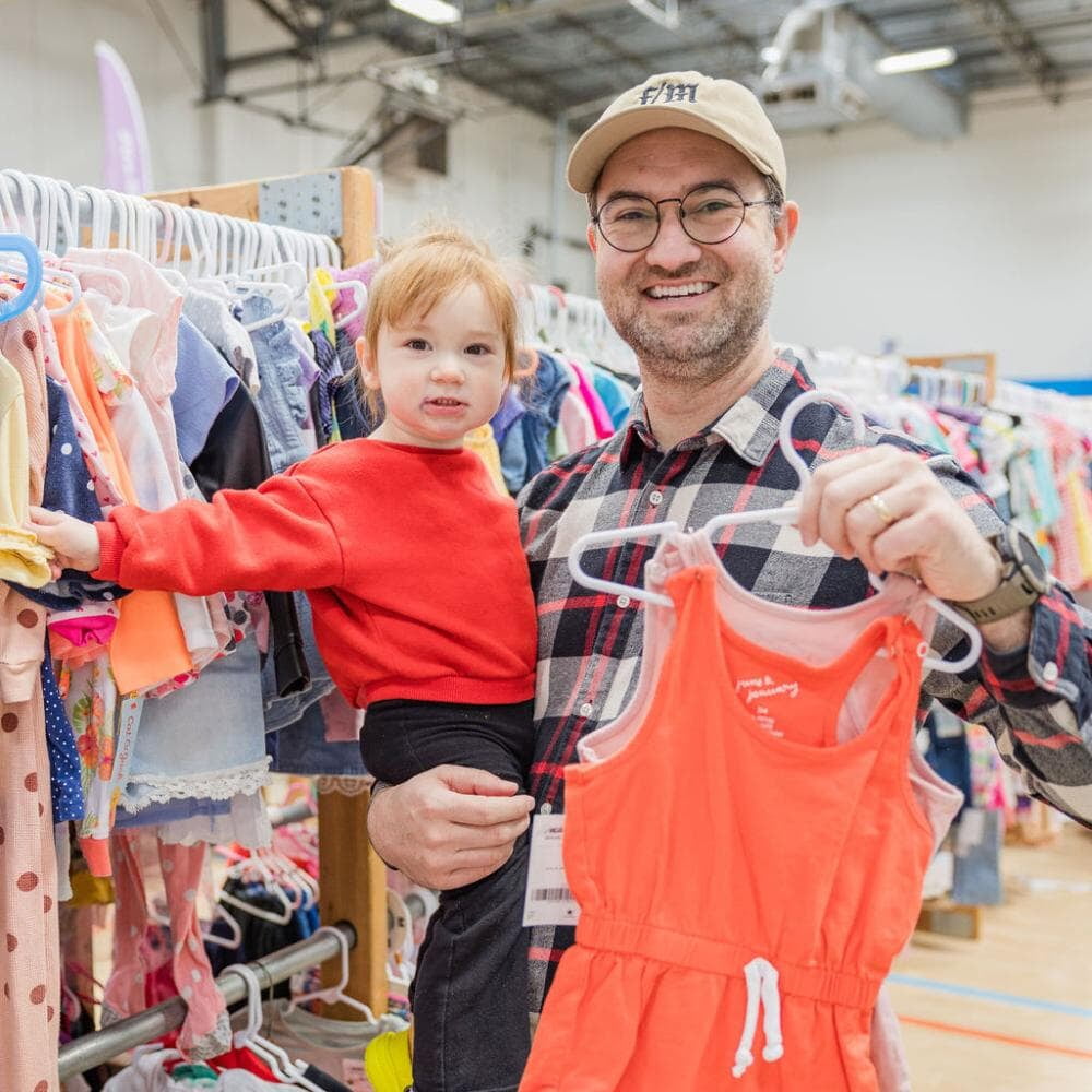 A dad smiles while holding toddler daughter with one arm and holding orange jumpsuit with the other hand.