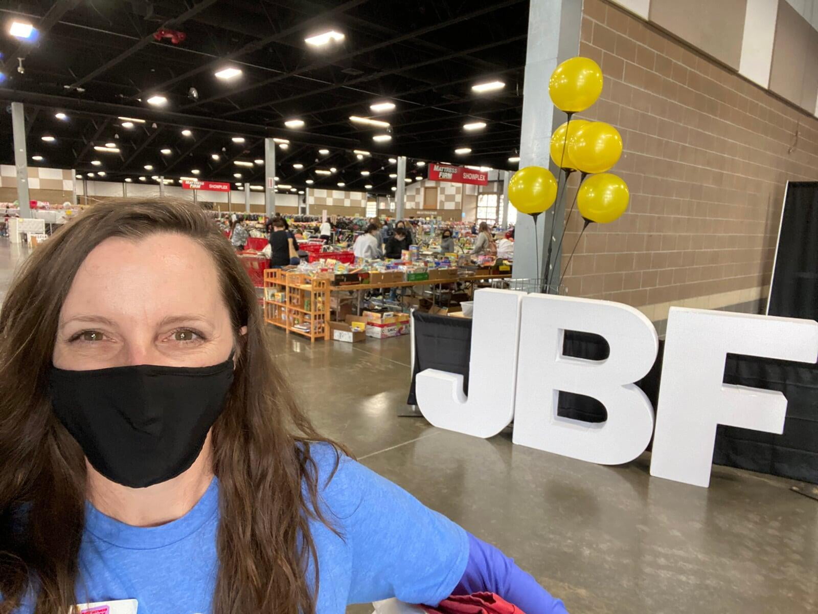 Owner/Operator, Allison Stephens, wears a face mask smiling at the cameral standing in front of the expansive sales floor with large JBF letters and yellow balloons in the background. Photo taken in 2020.