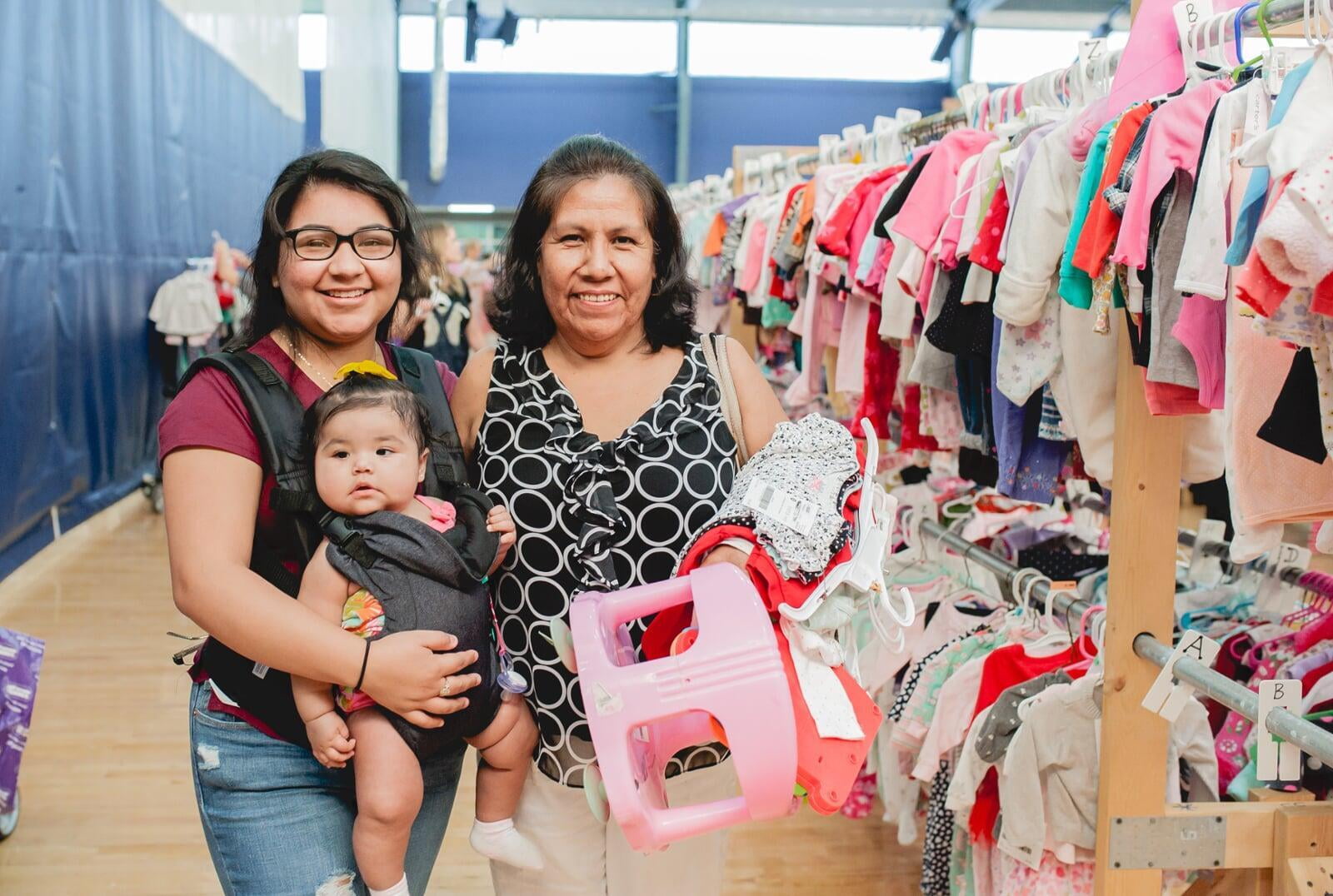 A mom, abuela and granddaughter in an infant carrier shopping for baby clothes. Abuela is holding a babyseat.