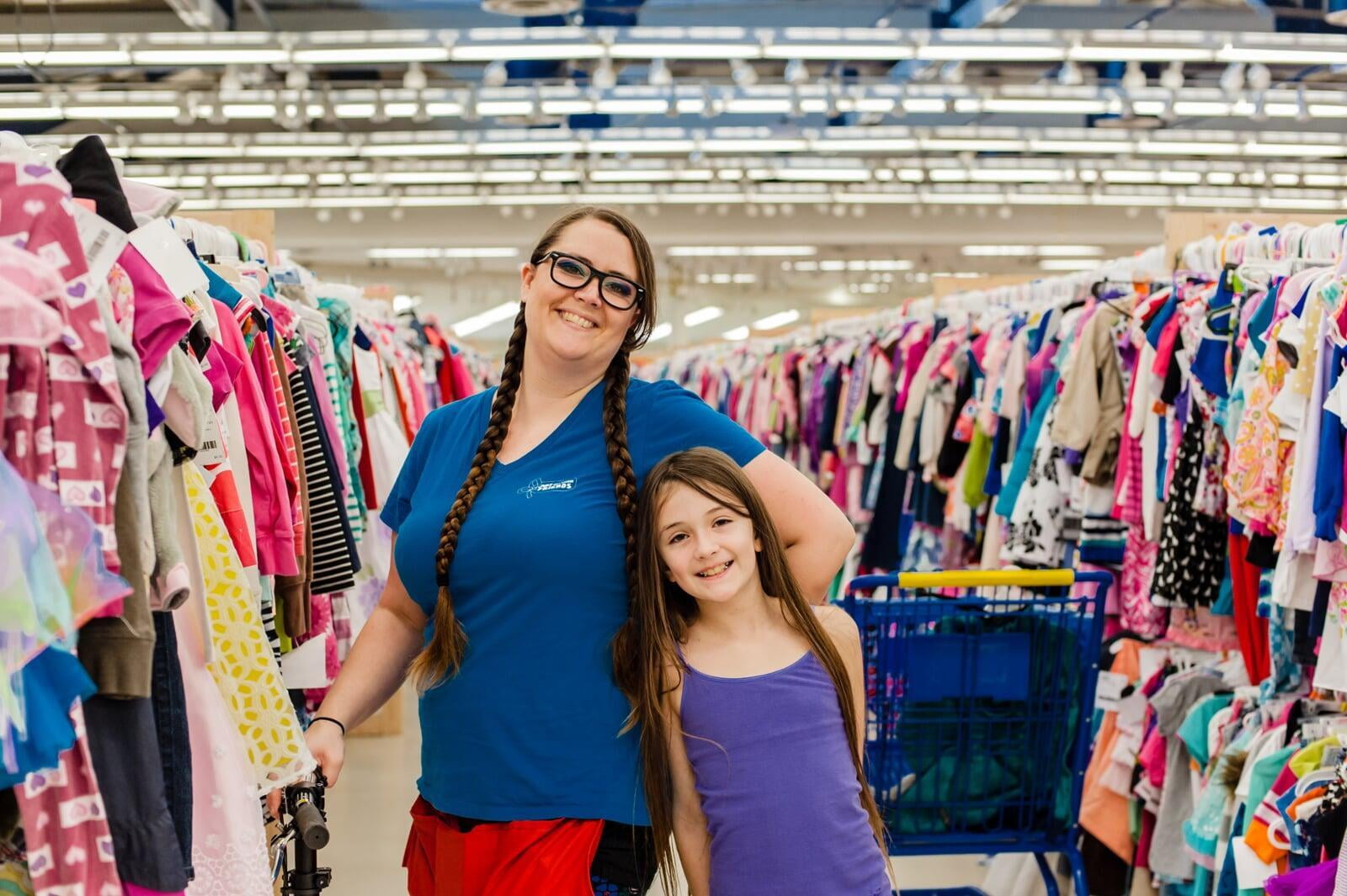 A mom and young daughter smile as they shop rows and rows of clothing.