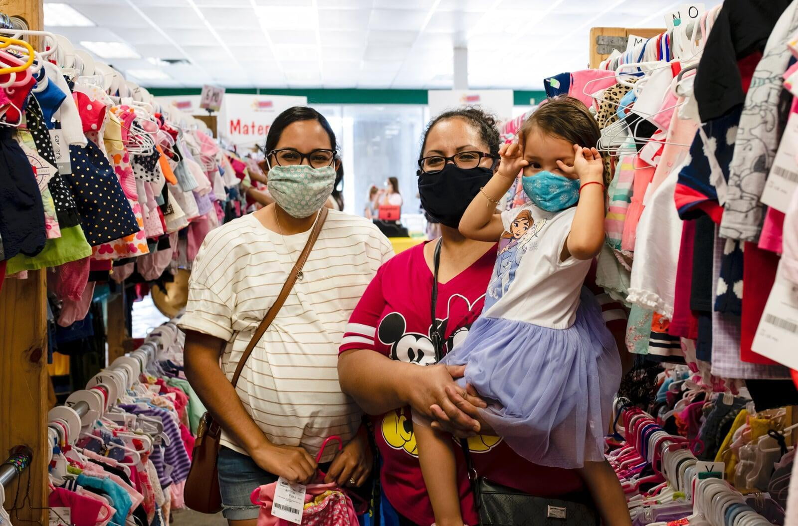 Two face masked moms—one holding a child with a mask, the other pregnant—shop for their families at the local JBF sale.