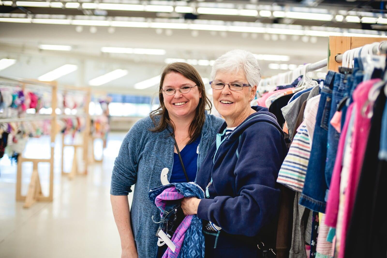 A mom and grandmother stand beside a long row of school age clothes for sale.