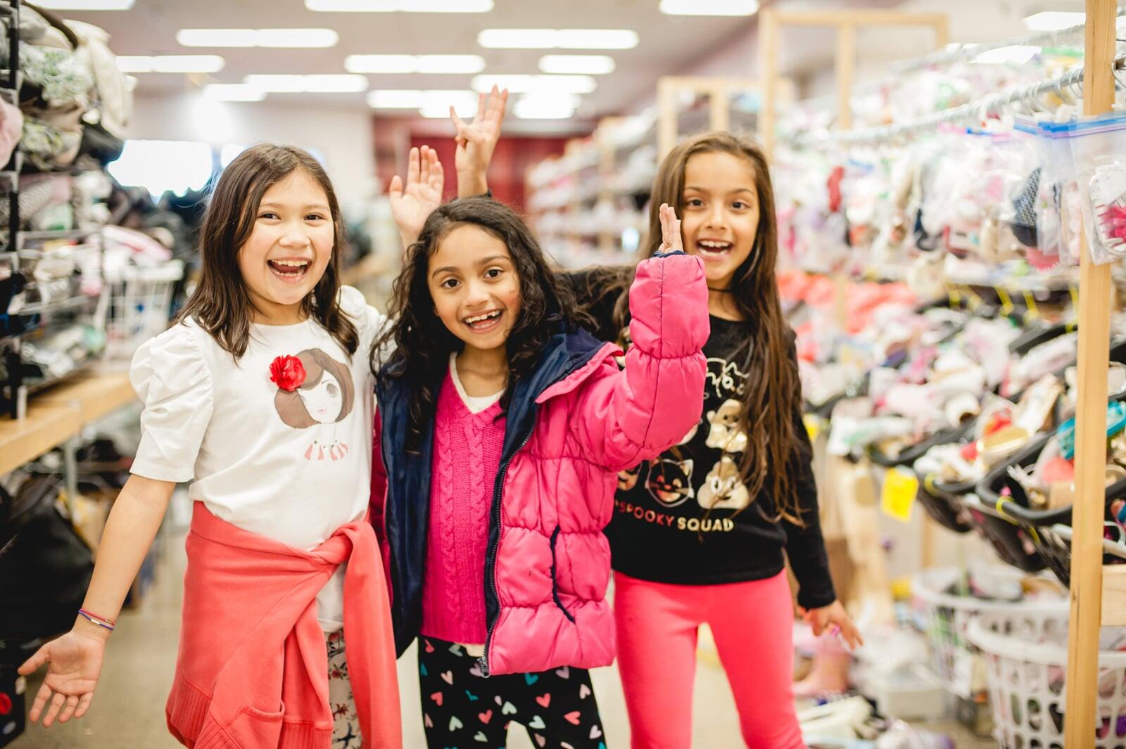 Three school aged girls smiling and holding hands in the air as they shop for shoes.